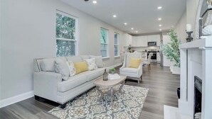 A modern living room with a sofa, armchair, and coffee table, a kitchen in the background, and a fireplace. - Blue Bungalow! Wonderful 4-bedroom house in charming Augusta (Augusta)