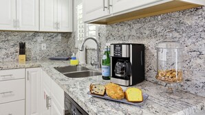 Una cocina con armarios blancos, encimera de granito, cafetera, un tarro de vidrio y una bandeja con pan.