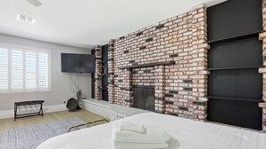 Un dormitorio con chimenea de ladrillo, una cama con ropa de cama blanca, un televisor de pantalla plana y una ventana con persianas.