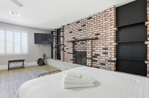 Ein Schlafzimmer mit Kamin aus Ziegelsteinen, einem Bett mit weißer Bettwäsche, einem Flachbildfernseher und einem Fenster mit Jalousien.