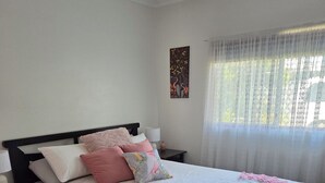 A neatly made bed with a white bedspread and pink accents, a bedside table with a lamp, and a window with sheer curtains. - Debs Place Room 2. Peaceful cosy room with your own entrance. (Denmark)
