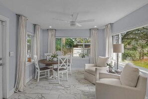Una habitación con una mesa de comedor, sillas, un sofá, un ventilador de techo y ventanas grandes con cortinas.