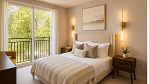 A bedroom with a large bed, two bedside tables, a nightstand, a chair, a window with a view of trees, and a painting on the wall. - Chevy Chase  301  2BR (Chevy Chase)