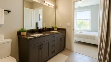A bathroom with a dark vanity, two sinks, a large mirror, and a view into a bedroom with a bed and window.