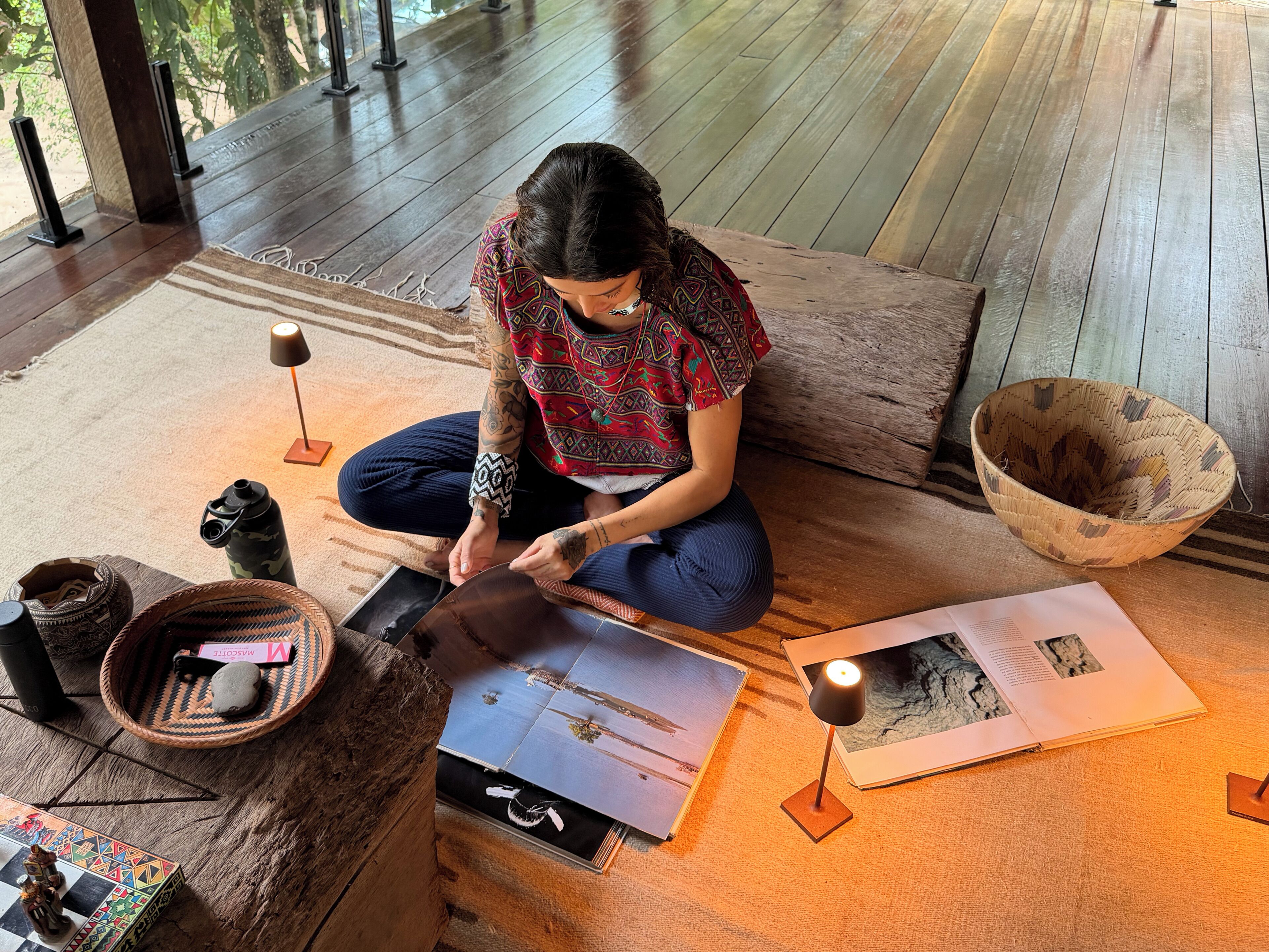 A person sitting on a wooden floor with a book, a lamp, and a woven basket.