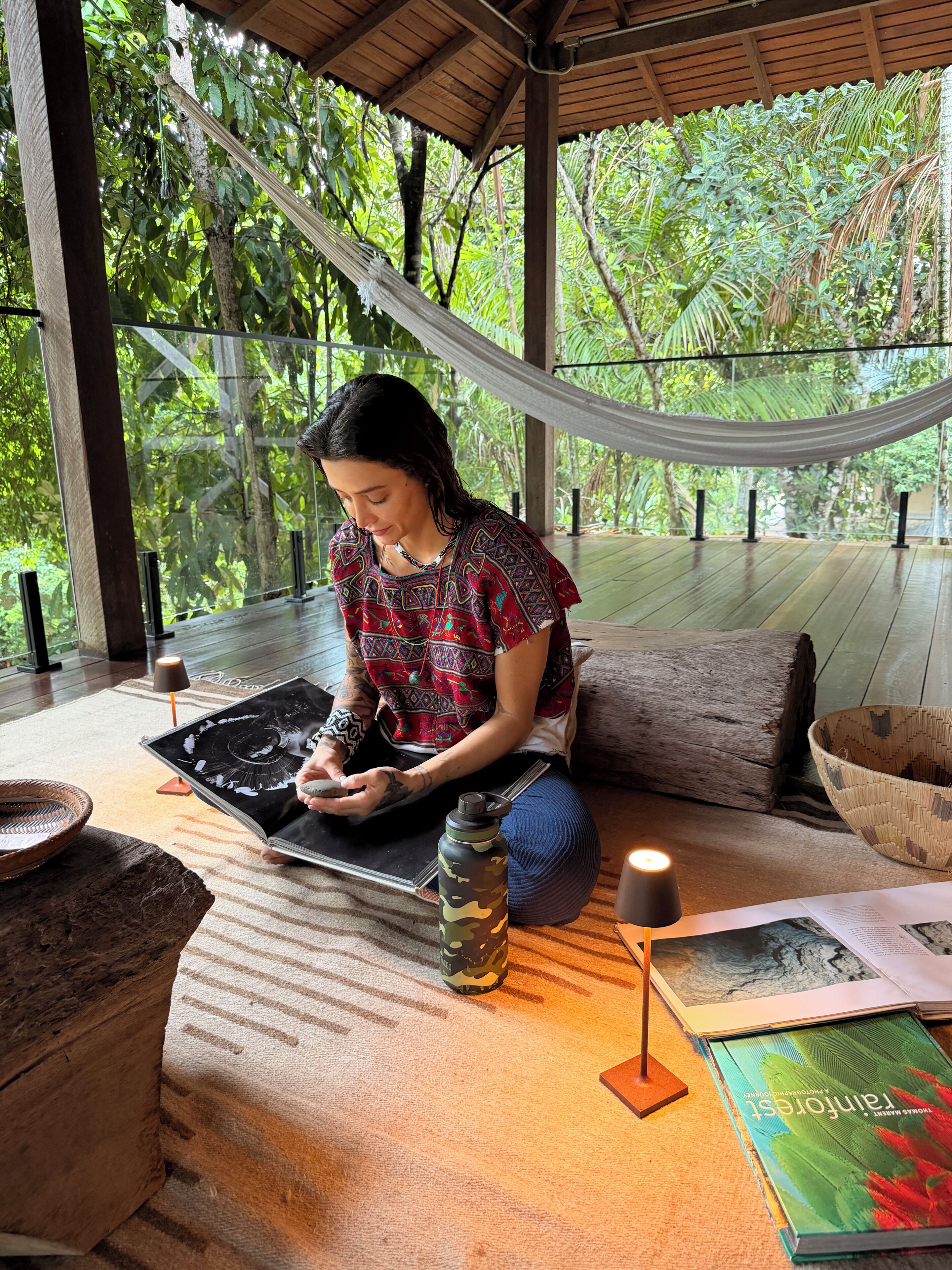 A person sitting on a hammock in a room with a wooden floor, surrounded by greenery, with a book and a water bottle on the floor.