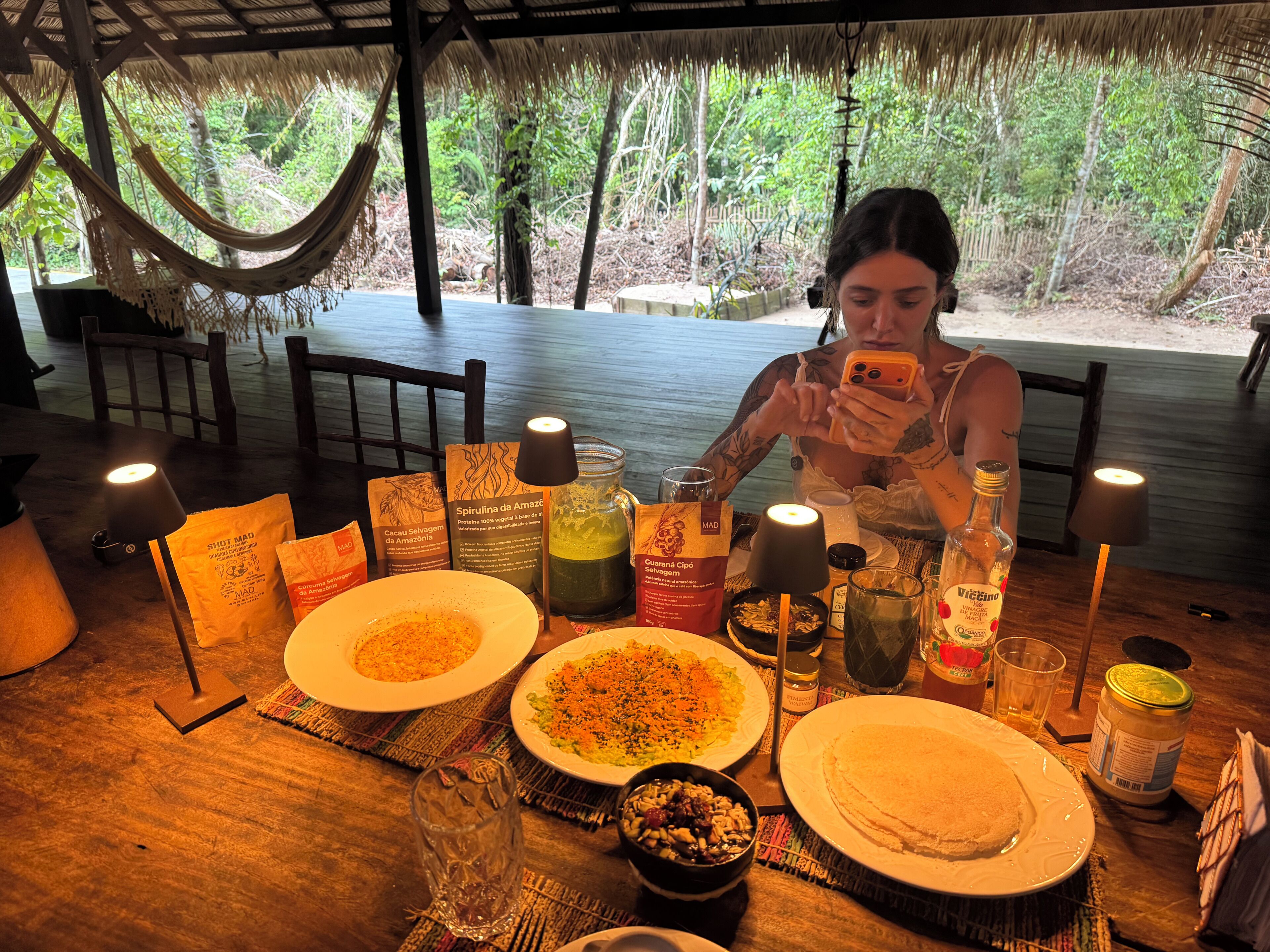A dining table with food, drinks, and candles in a thatched-roof area with a view of trees.