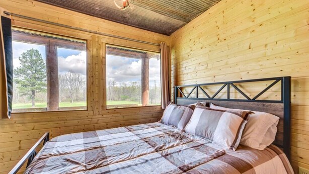 Un dormitorio de cabaña de madera con cama, ventanas amplias y vista a árboles.