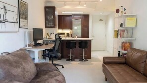 A modern living room with a desk, two bar stools, a brown leather sofa, a bookshelf, and a kitchen area with a refrigerator. - Quiet North Waterfront 1BR · WFH Setup · in-unit W/D · 24/7 Doorman, Gym, Sauna (San Francisco)