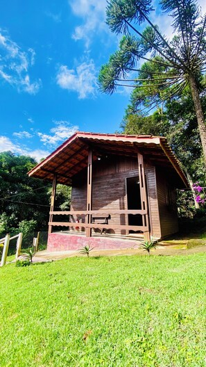 A wooden cabin with a porch, surrounded by grass and trees. - Pousada Domos Serra do Sol (Camanducaia)