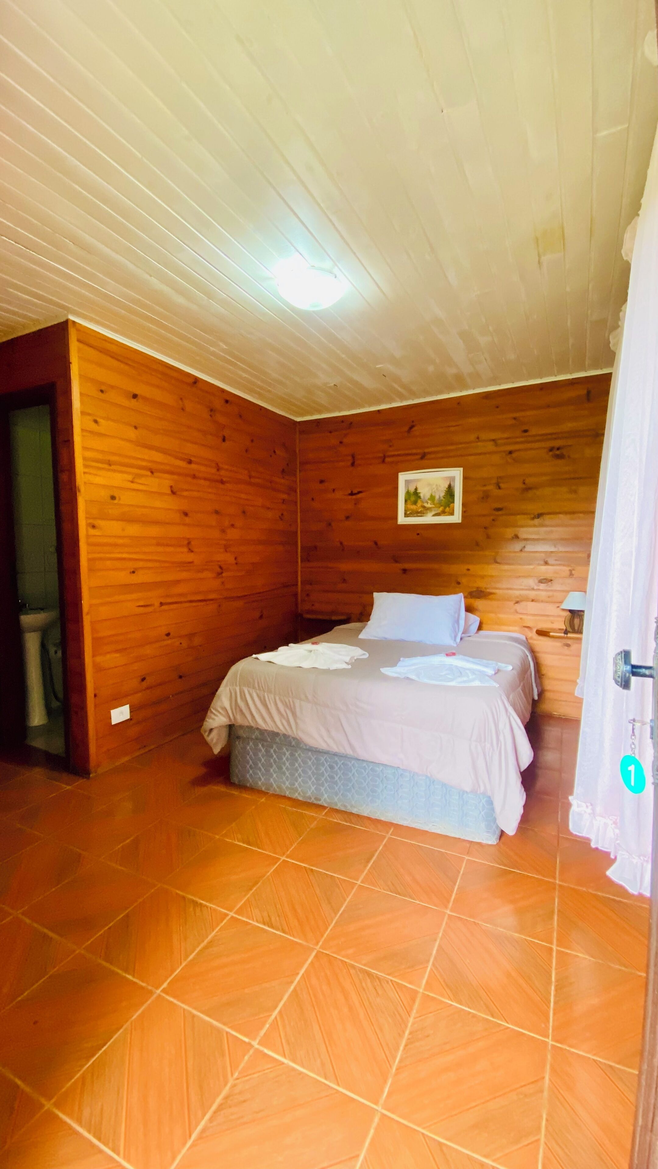 Una habitación con paredes de madera, una cama con ropa blanca, un cuadro en la pared y suelo de baldosas.