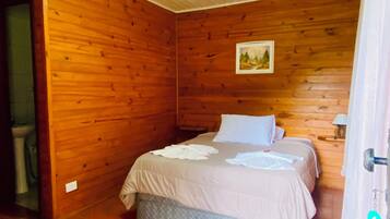 Una habitación con paredes de madera, una cama con ropa blanca, un cuadro en la pared y suelo de baldosas.