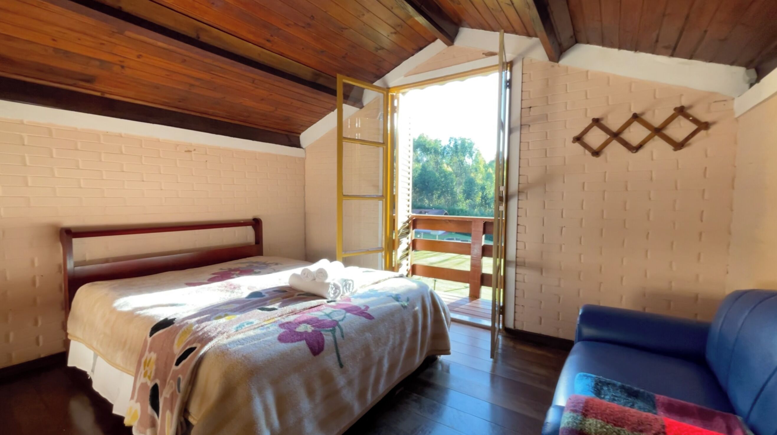 Un dormitorio con techo de madera, una cama con colcha estampada, un sofá azul y vistas a zonas verdes a través de una ventana.