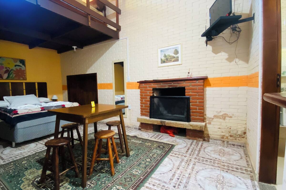 A room with a bed, a table, two stools, a fireplace, and a patterned rug.