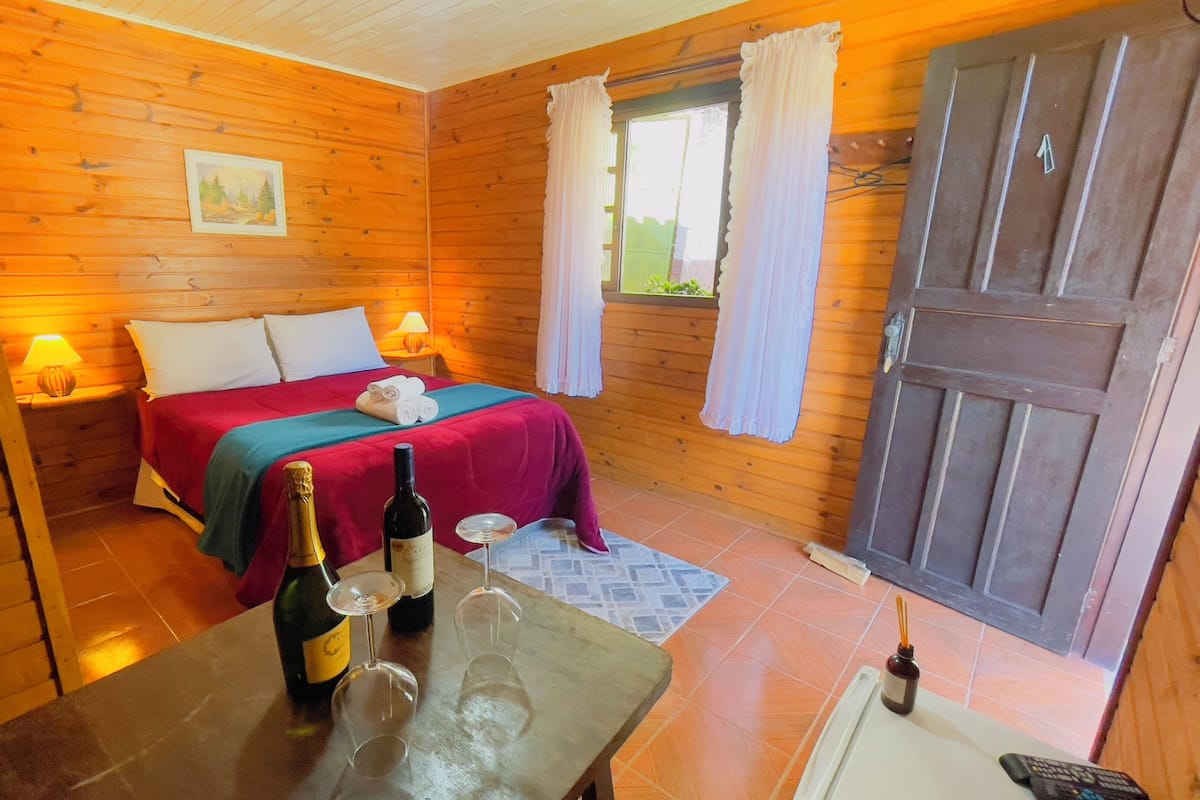 A wooden room with a bed, a table with a bottle and glasses, and a door.