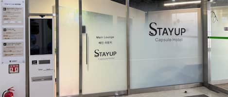 A Stayup Capsule Hotel entrance with a glass door, a vending machine, and a fire extinguisher.
