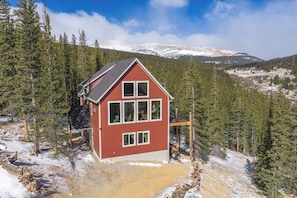 Une cabane rouge dotée d’un balcon, entourée de montagnes enneigées et d’une forêt dense.