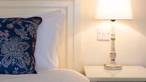 A bed with a floral pillow, a white bedside table with a lamp, and a wall switch. - Milsoms Hotel (Kenilworth)