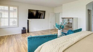 Een slaapkamer met een bed, een plafondventilator, een aan de muur bevestigde televisie en een raam met jaloezieën.