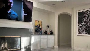 Een moderne woonkamer met een flatscreen televisie, een open haard, een gang naar een andere kamer en een groot abstract schilderij aan de muur.