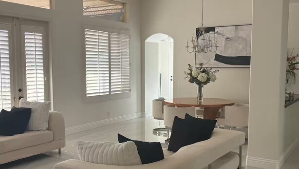 Een ruime woonkamer met een hoge plafond, grote ramen met zonwering, een eethoek met een ronde tafel en een kroonluchter.