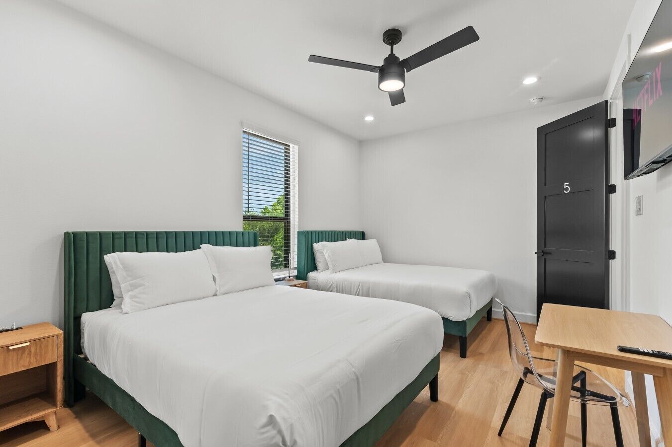 Ein Zimmer mit zwei Betten, einem Schreibtisch, einem Stuhl, einem Deckenventilator und einem Fenster mit Jalousien.