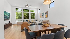 Un soggiorno moderno con divano, tavolo da pranzo e sedie, ventilatore a soffitto e ampie finestre con vista sul verde.