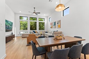 A modern living room with a sofa set, a dining table with chairs, a ceiling fan, and large windows with a view of greenery. - Luxury 7BR/7BA Home • Skyline Game Room • Houston (Houston)