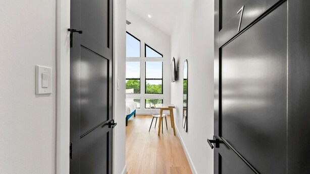 Un corridoio moderno con porte scure, una sala da pranzo con tavolo e sedie, e ampie finestre che offrono vista sul verde.