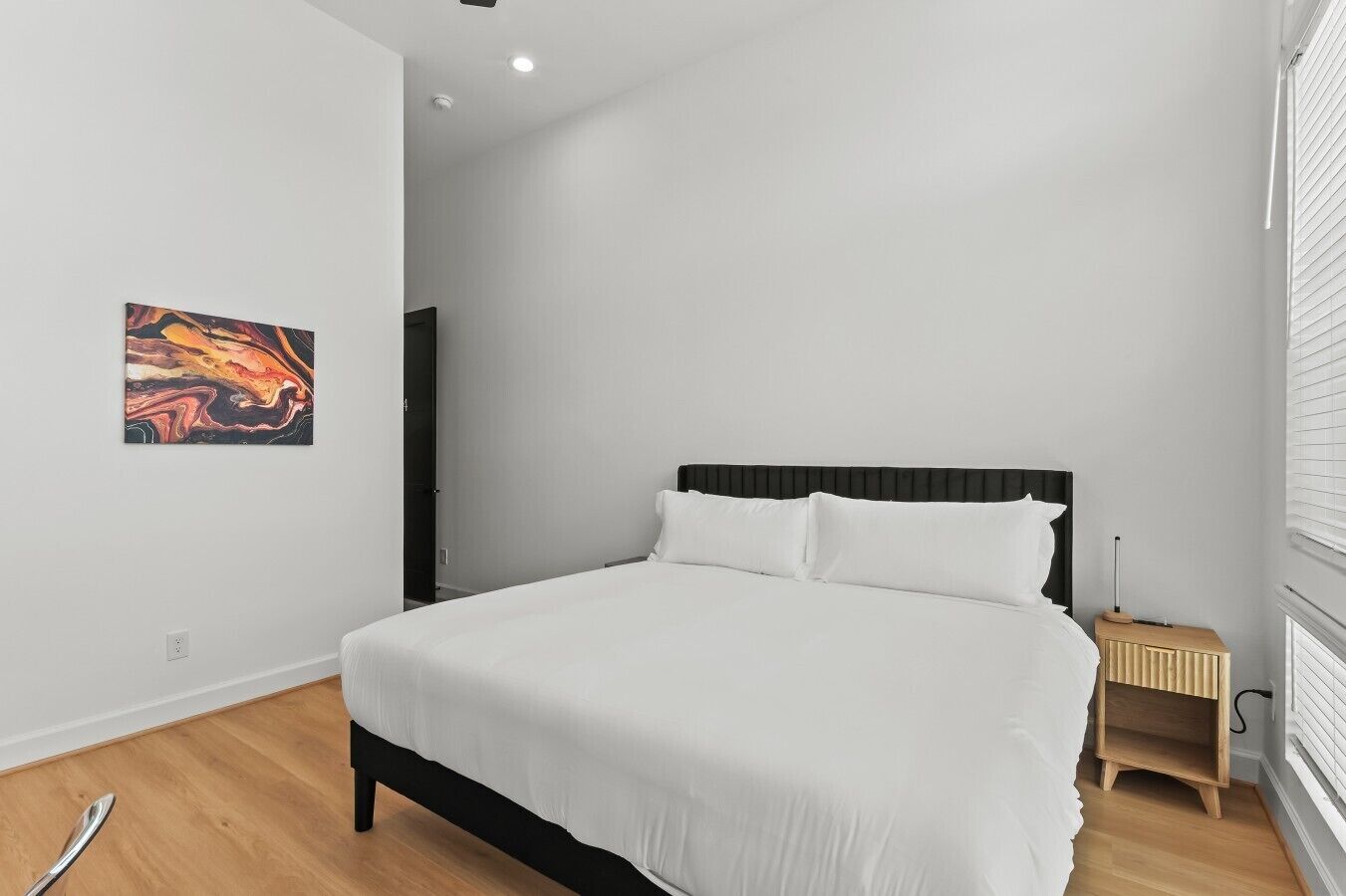 A bedroom with a bed, a bedside table, a wall-mounted artwork, and a window with blinds.
