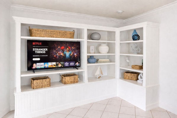 A white entertainment center with a flat-screen TV displaying a Netflix show, shelves with decorative items, and woven baskets.