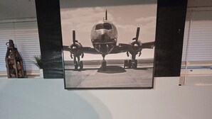 Une pièce avec une table et deux chaises, un tableau encadré représentant un avion accroché au mur, et une fenêtre avec des stores.