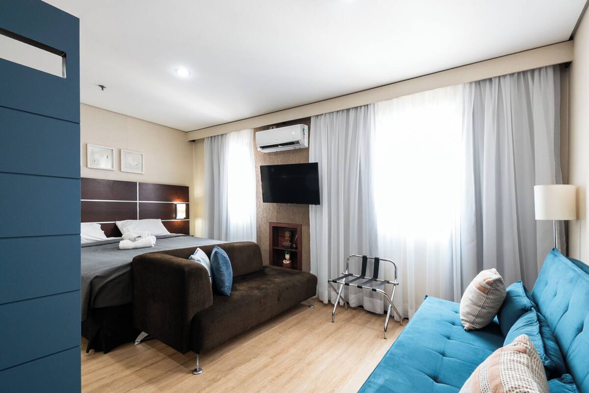 A hotel room with a bed, two sofas, a TV, and an air conditioning unit.