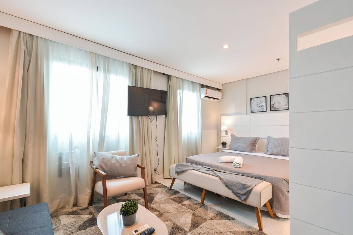 A modern hotel room with a bed, a TV, a sofa, a coffee table with a plant, and a large window with curtains.