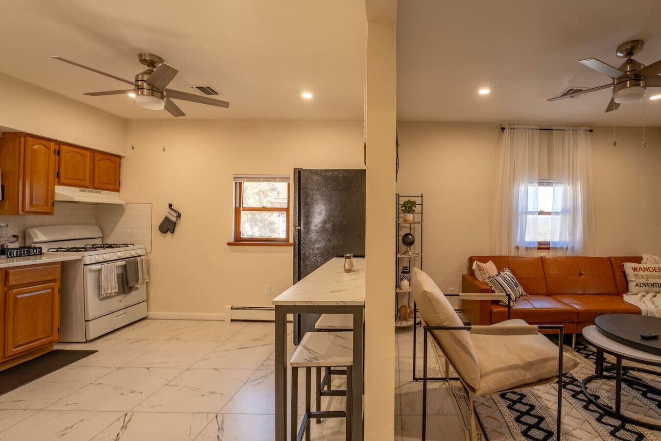 A kitchen with wooden cabinets, a stove, and a dining area with a table and chairs. There are ceiling fans, a window, and a sofa in the living area.