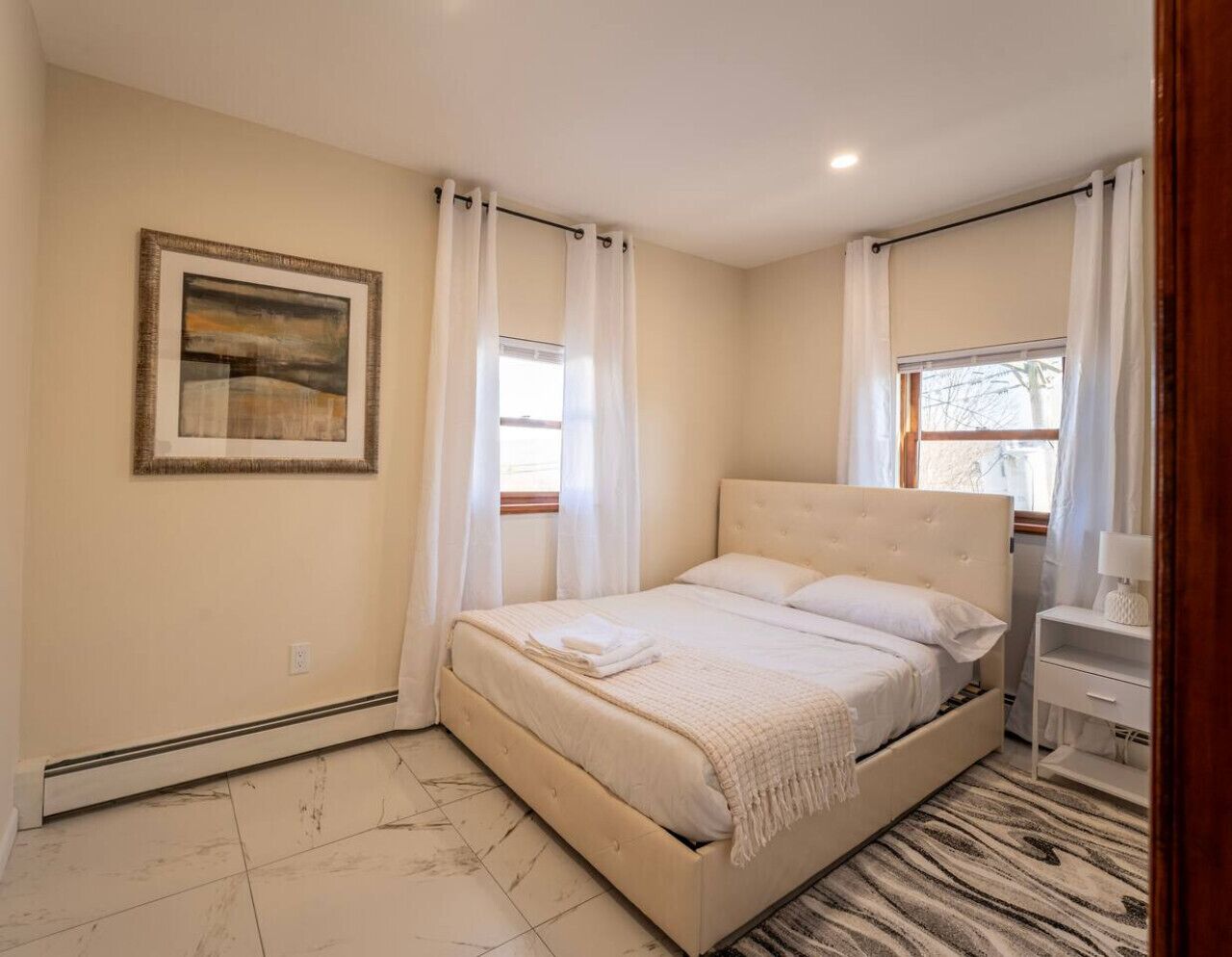 A bedroom with a bed, a bedside table, a nightstand, a window, and a painting on the wall.