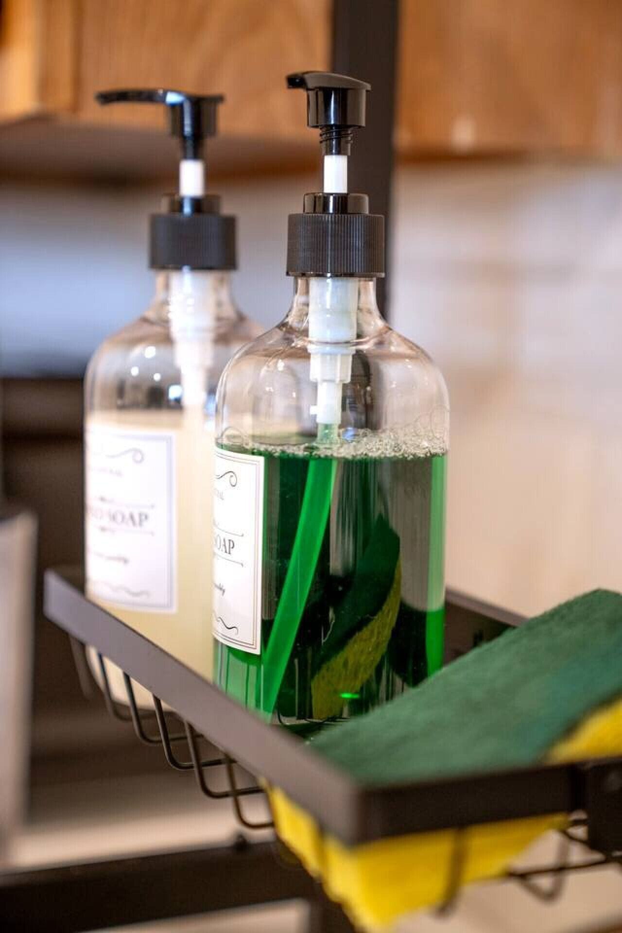 Two bottles of liquid soap in a metal rack with a green cleaning cloth.