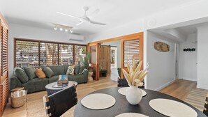 A modern living room with a sofa, coffee table, and dining table. There is a ceiling fan, a window with blinds, and a door leading to another room. - Absolute Beachfront | Palm Cove Apt Sleeps 4 (Palm Cove)