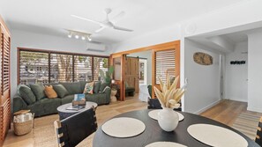 Ein modernes Wohnzimmer mit Sofa, Couchtisch und Esstisch. Es gibt einen Deckenventilator, ein Fenster mit Jalousien und eine Tür zu einem anderen Raum.