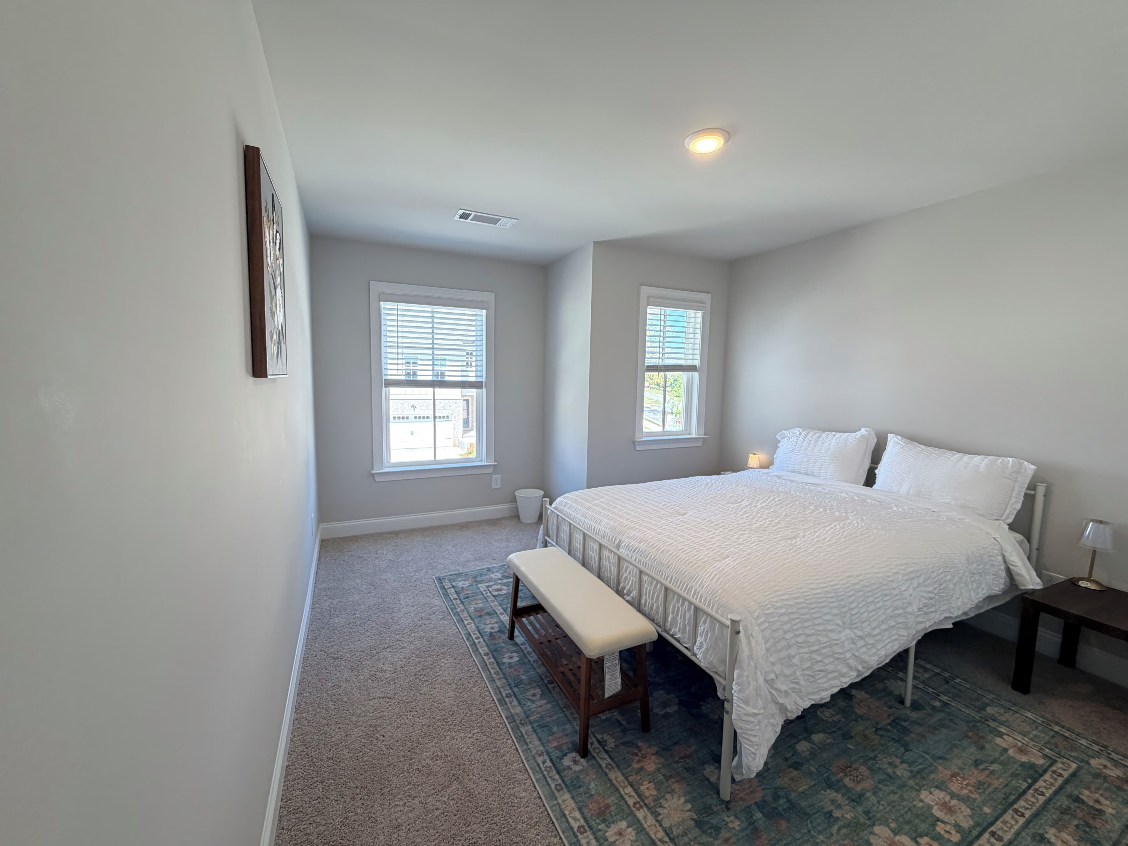 A bedroom with a large bed, two windows, a bench, a bedside table, and a floor lamp.