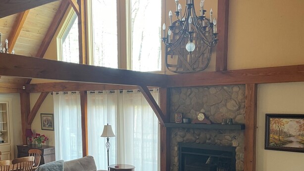 A spacious living room with a stone fireplace, wooden beams, a chandelier, and a large window.