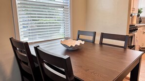 A dining table with a decorative bowl, four chairs, a window with blinds, and a kitchen area with cabinets and appliances. - Historic Downtown Visalia 2BR • 1BA (Visalia)
