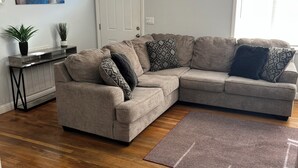 Un salon comprenant un canapé d’angle, une table basse, une plante en pot, un tableau accroché au mur et une porte avec une fenêtre.