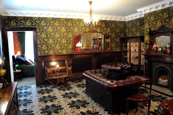A room with a patterned wallpaper, a chandelier, a leather sofa, a wooden cabinet, and a fireplace.