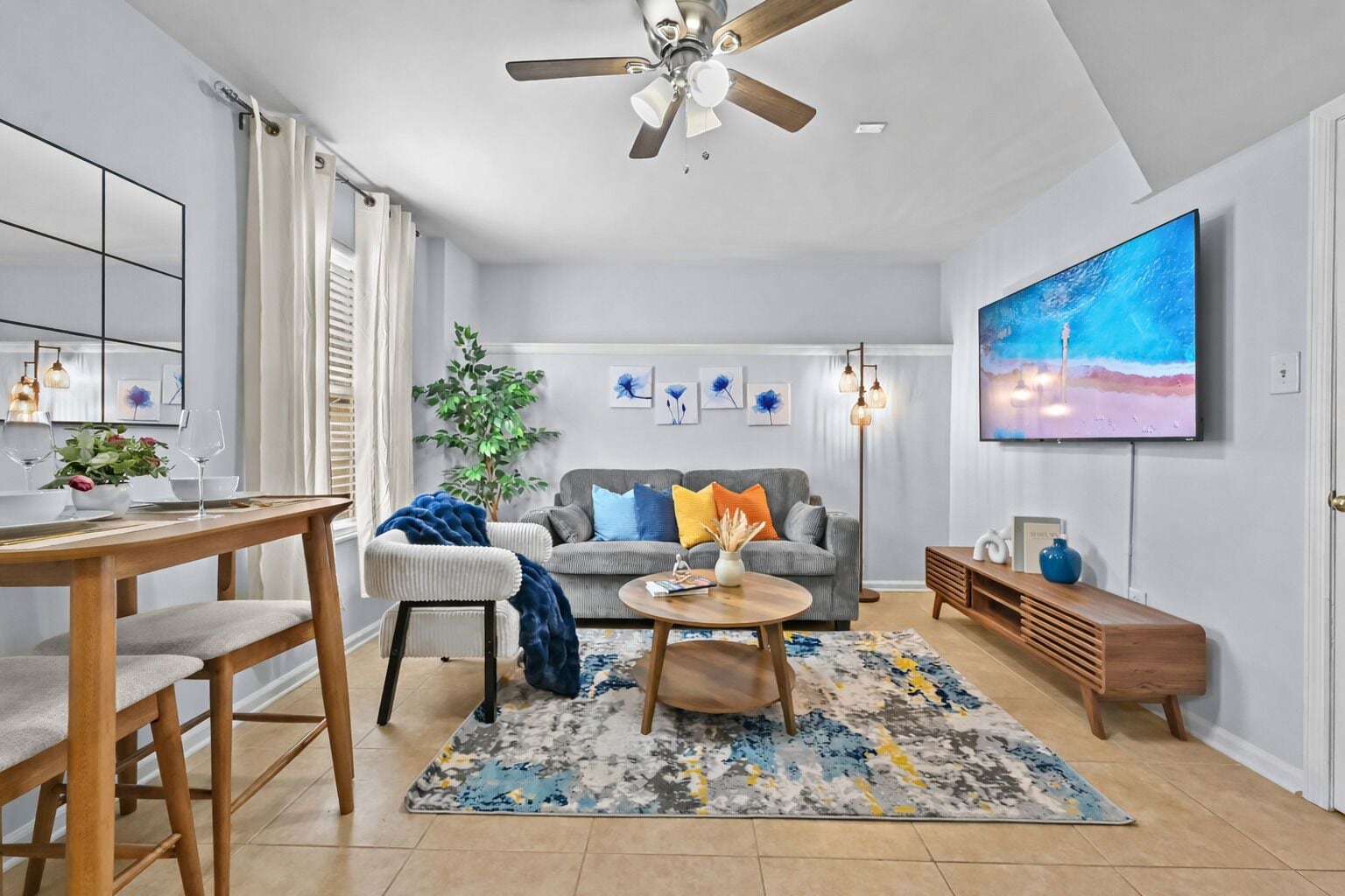 A modern living room with a dining area, a ceiling fan, a flat-screen TV, and a large painting.