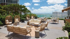 An outdoor patio with wicker furniture, a sofa, and tables, overlooking a body of water and city skyline. - LUXORA 2 BR HIGH FLOOR W RESIDENCES-FREE SPA (Miami)