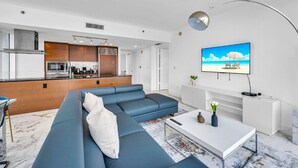A modern living room with a blue sofa, a white coffee table, and a kitchen area with wooden cabinets and a stainless steel refrigerator. - LUXORA 2 BR HIGH FLOOR W RESIDENCES-FREE SPA (Miami)