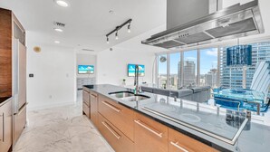 A modern kitchen with a large island, stainless steel appliances, and a city view through large windows. - LUXORA 2 BR HIGH FLOOR W RESIDENCES-FREE SPA (Miami)
