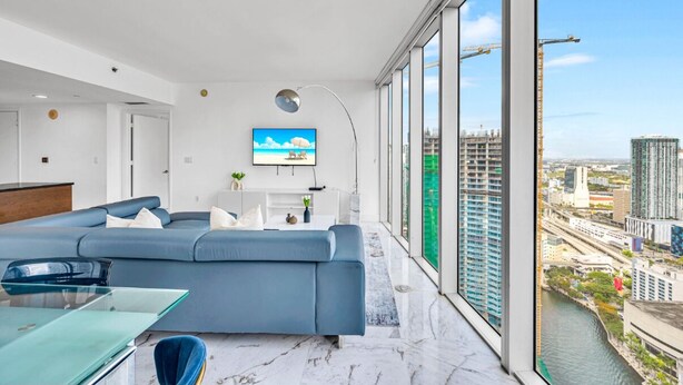 A modern living room with a large sofa, a glass coffee table, a flat-screen TV, and a view of a cityscape through large windows. - LUXORA 2 BR HIGH FLOOR W RESIDENCES-FREE SPA (Miami)