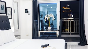 A hotel room with a bed, a desk with a potted orchid, a chair, a window with curtains, and a balcony with a railing. - Kandy Haven - Studio Type Unit (Angeles City)